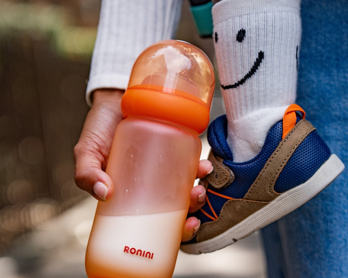 A parent holding The Wrapped Glass Bottle next to a toddler’s sneaker